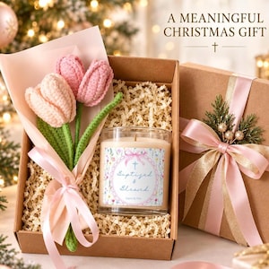 Puede incluir: Una caja de regalo que contiene un ramo de ganchillo de dos tulipanes rosas y una vela de cristal con una etiqueta que dice A MEANINGFUL CHRISTMAS GIFT y Baptized & Blessed. La caja está llena de papel picado e incluye un lazo rosa.
