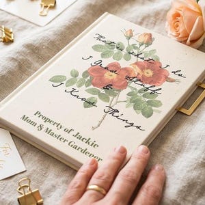 May include: Cream-colored journal with a floral design featuring red and orange roses and green leaves. The journal has gold accents, a gold bookmark, and the text "That's what I do, I Grow Stuff & I Know Things." The journal is labeled "Property of Jackie, Mom & Master Gardener."