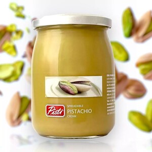 May include: A jar of pistachio cream with a silver lid and a label that reads "Pisti Pistachio Cream." The cream is a light green colour, and the label features an illustration of a pistachio nut. Pistachios are scattered around the jar.