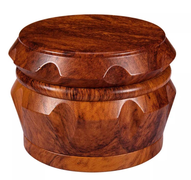 May include: A wooden grinder with a rich, dark brown finish. The grinder has a round, flat top and a cylindrical base with a series of angled, carved facets. The wood grain is visible throughout the design.
