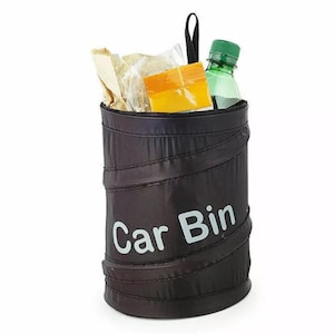 May include: Black car bin with the words "Car Bin" in white. The bin is cylindrical and contains a green water bottle, a yellow snack, and some paper. A black handle is attached to the top.