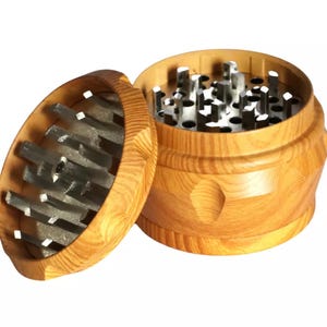 May include: A wooden herb grinder with a light brown finish. The grinder is cylindrical with a lid and a base. The lid is open, revealing the metal grinding teeth. The base has a series of decorative indentations.