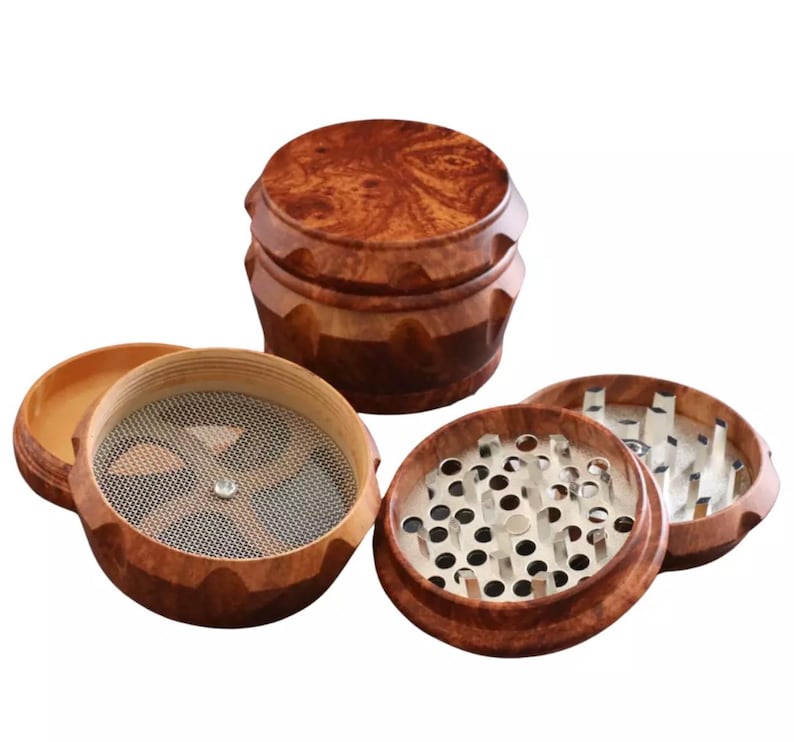 May include: A wooden herb grinder with multiple disassembled parts. The grinder is made of a dark brown wood with a natural grain pattern. The components include a top, a grinding chamber with metal teeth, a mesh screen, and a bottom chamber.