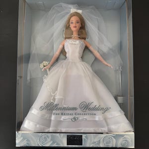 Peut inclure: Poupée blonde vêtue d'une robe de mariée blanche, d'un voile et d'un collier de perles, présentée dans une boîte en plastique transparente. La boîte indique "Millennium Wedding" et "The Bridal Collection". L'emballage est décoré d'un motif floral.