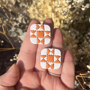 May include: Two square earrings with a white base and an orange starburst pattern. The earrings have rounded corners and are held in a hand. The background is blurred with dried flowers.