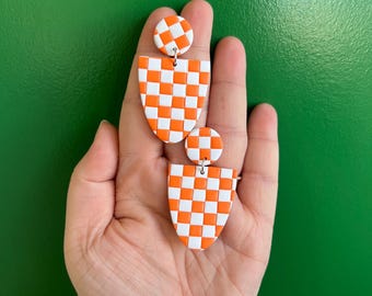 May include: A pair of orange and white checkered earrings. Each earring has a small circular stud and a larger shield-shaped drop. The earrings are held in a hand against a green background. The earrings are made of a lightweight material.