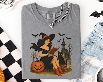 May include: Gray Comfort Colors t-shirt featuring a Halloween-themed illustration. The design includes a witch reading a book, a jack-o'-lantern, a black cat, and a haunted house. Bats fly in the background, creating a spooky atmosphere.