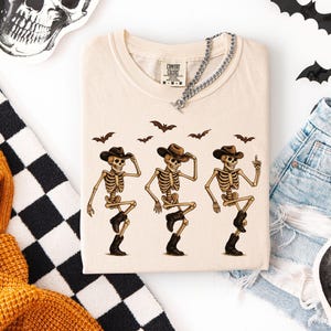 May include: Cream-colored t-shirt featuring a graphic of three dancing skeletons wearing cowboy hats and boots. Brown bats are above the skeletons. A silver chain necklace is draped on the shirt. Halloween-themed apparel.