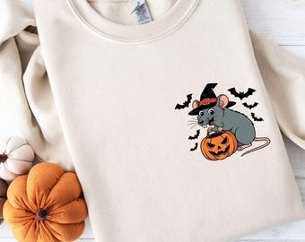 May include: A cream-colored sweatshirt featuring a Halloween-themed graphic. The graphic depicts a gray rat wearing a witch hat and holding a carved orange pumpkin. Black bats are scattered around the rat. The sweatshirt is folded on a white surface.