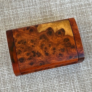 Könnte beinhalten: Rechteckige Holzbox mit einem reichen, wirbelnden Maserungsmuster in Braun- und Goldtönen. Die Box hat eine glatte, polierte Oberfläche und einen dunkelbraunen Rahmen. Die Holzmaserung weist dunkle Knoten und komplizierte Muster auf.