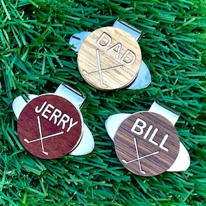 May include: Three personalized golf ball markers with a silver clip. The markers are made of wood and feature the names "DAD", "JERRY", and "BILL" with a crossed golf club design.