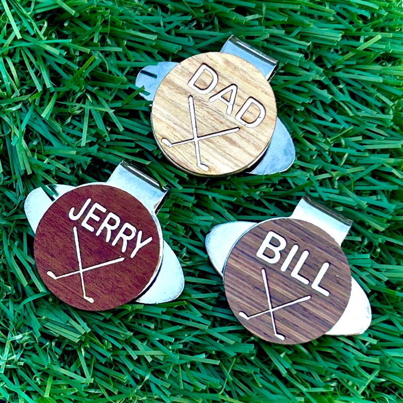 May include: Three personalized golf ball markers with a silver clip. The markers are made of wood and feature the names "DAD", "JERRY", and "BILL" with a crossed golf club design.