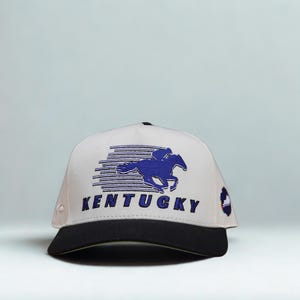 May include: A white baseball cap with a black brim featuring an embroidered blue graphic of a horse and jockey in motion above the word KENTUCKY in blue block letters. A small circular patch with a blue and white design is on the side panel.