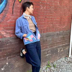 Może przedstawiać: Osoba ubrana w niebieską kurtkę dżinsową z kieszeniami w stylu patchwork, beżową koszulę i ciemnoniebieskie dżinsy. Osoba opiera się o czerwoną ceglaną ścianę z graffiti. Czarno-białe trampki dopełniają strój.