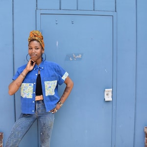 May include: A person wearing a blue short-sleeved button-down shirt with floral print pockets and cuffs, paired with blue jeans. The person is wearing a black top, a brown belt, and a patterned head wrap. The background is a blue door.