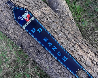 May include: A dark blue leather rifle sling with the text "DOUBLE D RANCH" and a Texas flag design with a steer skull. The sling has blue stitching and a black leather strap.