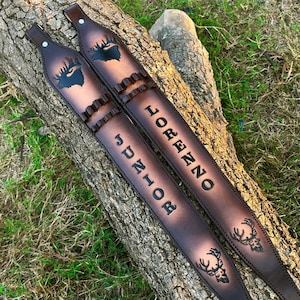 May include: Two brown leather rifle slings with embossed deer designs and the names "Lorenzo Junior". The slings have decorative stitching and metal rivets. They are lying on a tree trunk.