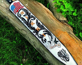 May include: A white leather rifle sling with black and orange decorative elements. The design includes an American flag, the letters "N" and "K", and a deer head. The sling has a black leather strap.