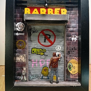 Puede incluir: Diorama en miniatura enmarcado de una barbería con arte de graffiti. La palabra "BARBER" está en letras amarillas sobre una puerta de garaje gris. Se muestra una figura pintando con aerosol la puerta. La puerta tiene un letrero de "No Parking".