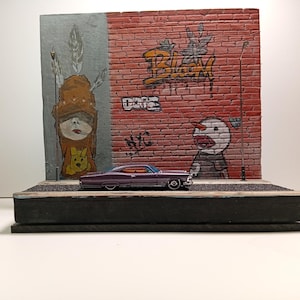 Puede incluir: Un diorama en miniatura que presenta un coche clásico morado estacionado frente a una pared de ladrillo con arte de graffiti. El arte mural incluye una figura nativa americana y un personaje parecido a un muñeco de nieve. El diorama está sobre una base gris oscuro.