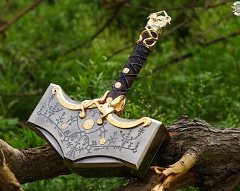 May include: A detailed, metallic hammer with gold accents and intricate engravings. The handle is wrapped in dark leather with gold detailing. The hammer is resting on a tree branch against a blurred green background.