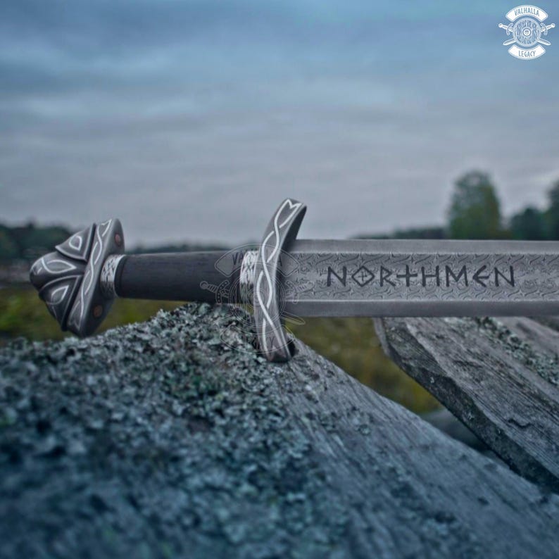 May include: A detailed image of a Viking sword with the word "NORTHMEN" etched into the blade. The sword features intricate silver-colored designs on the hilt and guard, with a dark handle. The sword rests on a weathered, gray stone surface.