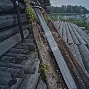 May include: A long, silver sword with an ornate handle and blade, resting on weathered wooden planks. A black scabbard leans against a log wall. The scene evokes a historical or fantasy theme, with a lake in the background.