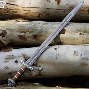 May include: A detailed image of a long sword with a patterned blade and a wooden handle. The sword's blade has a textured, wavy pattern. The handle is brown with silver accents. The sword is resting on a pile of logs.