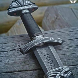 May include: A detailed close-up of a Viking sword. The sword features a dark wooden handle, a silver crossguard, and a blade with etched patterns and the word "NOK". The sword is set against a wooden background.