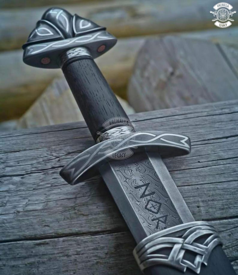 May include: A detailed close-up of a Viking sword. The sword features a dark wooden handle, a silver crossguard, and a blade with etched patterns and the word "NOK". The sword is set against a wooden background.