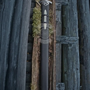 May include: A black sword in a black sheath with silver accents, resting against a wooden background. The sword features a detailed hilt and a decorative design on the sheath. The Valhalla Legacy logo is visible in the upper right corner.