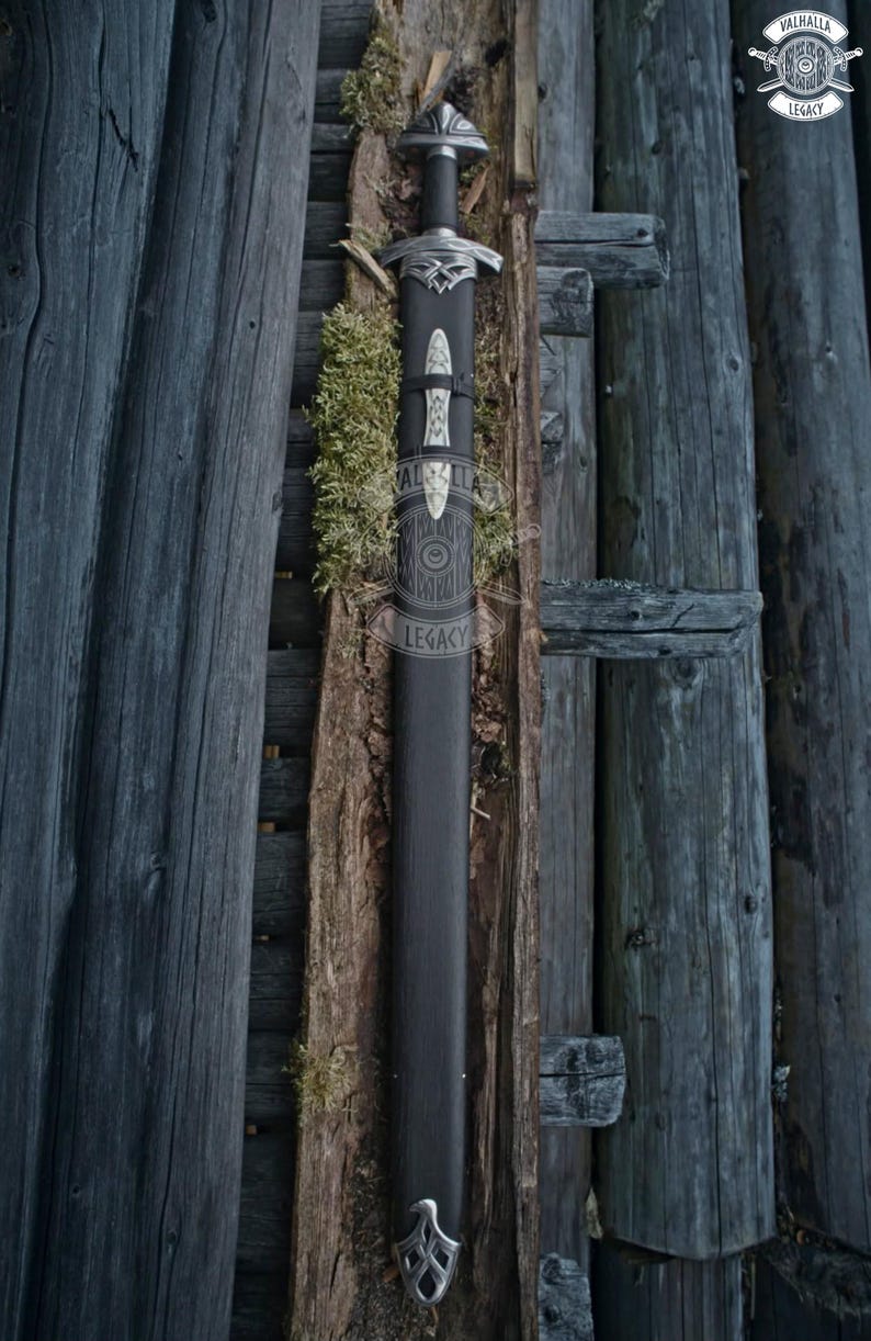 May include: A black sword in a black sheath with silver accents, resting against a wooden background. The sword features a detailed hilt and a decorative design on the sheath. The Valhalla Legacy logo is visible in the upper right corner.