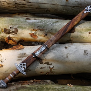 May include: A wooden sword with a brown handle and sheath, accented with silver-colored metal fittings. The blade is not visible. The sword rests on a pile of light-colored logs. The sword's design suggests a historical or fantasy theme.