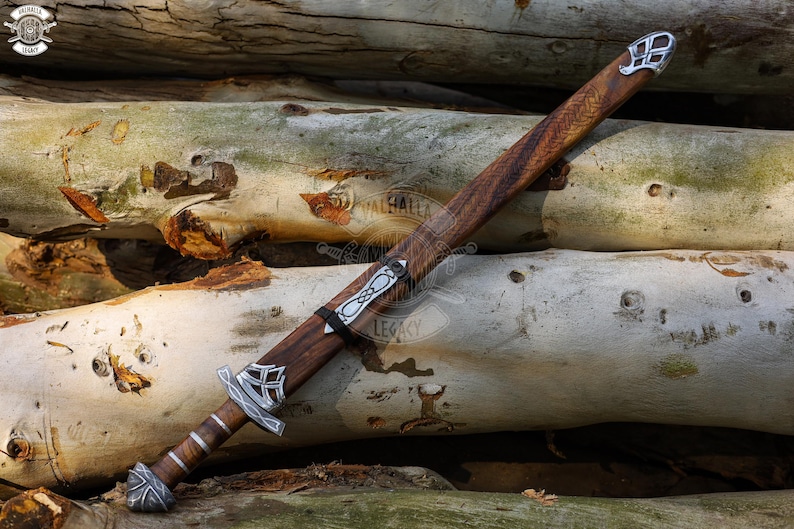 May include: A wooden sword with a brown handle and sheath, accented with silver-colored metal fittings. The blade is not visible. The sword rests on a pile of light-colored logs. The sword's design suggests a historical or fantasy theme.