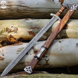 May include: A detailed image of a sword and scabbard. The sword has a patterned blade and a wooden handle with silver accents. The scabbard is made of wood with intricate carvings and silver details. The sword and scabbard are resting on a bed of logs.