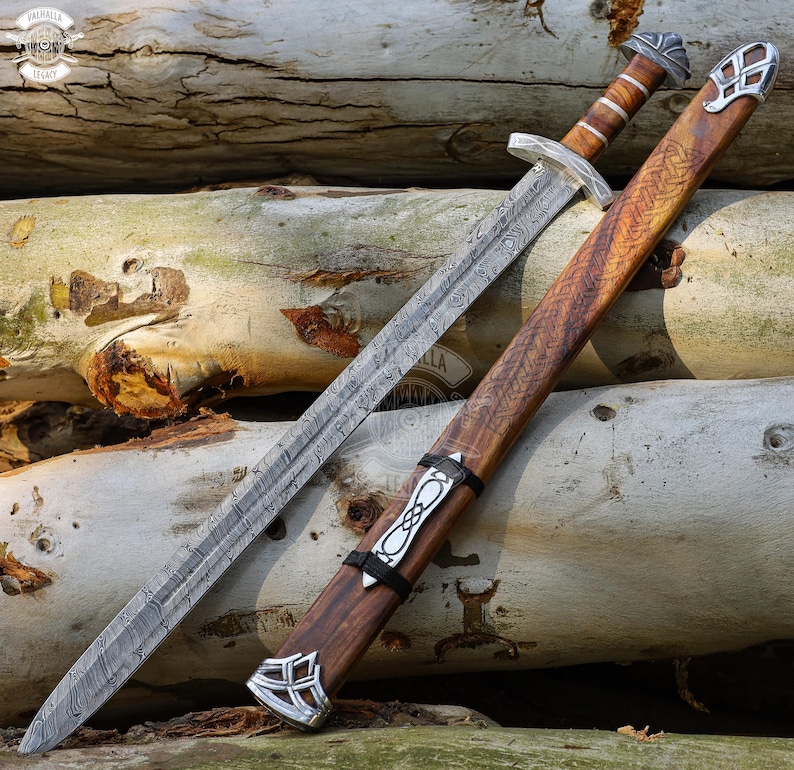 May include: A detailed image of a sword and scabbard. The sword has a patterned blade and a wooden handle with silver accents. The scabbard is made of wood with intricate carvings and silver details. The sword and scabbard are resting on a bed of logs.