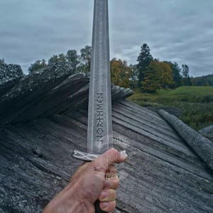 May include: A long, silver sword with the word "NORTHMEN" etched on the blade. The sword has a detailed hilt and is held against a backdrop of weathered wood and a cloudy sky. The Valhalla Legacy logo is in the upper right corner.