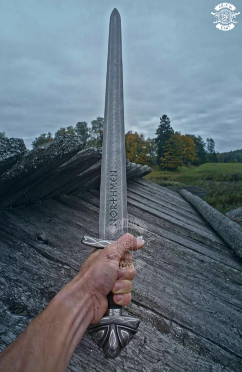 May include: A long, silver sword with the word "NORTHMEN" etched on the blade. The sword has a detailed hilt and is held against a backdrop of weathered wood and a cloudy sky. The Valhalla Legacy logo is in the upper right corner.