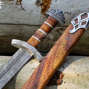 May include: A detailed image of a Viking sword with a patterned blade and wooden handle. The handle is wrapped with silver bands. The wooden sheath has an engraved Celtic knot design and silver accents. The sword is displayed on a wooden surface.