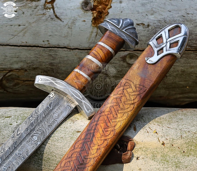 May include: A detailed image of a Viking sword with a patterned blade and wooden handle. The handle is wrapped with silver bands. The wooden sheath has an engraved Celtic knot design and silver accents. The sword is displayed on a wooden surface.