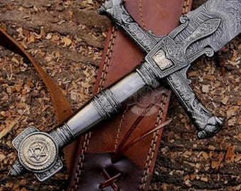May include: An ornate, silver-colored sword with a detailed handle and crossguard, resting on a brown leather sheath. The blade features a unique pattern. The sword's handle has a gold-colored emblem. The sheath is secured with a leather strap.