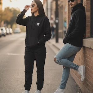 May include: Two people wearing black hoodies with the text "SAS" on the chest, black pants, and white sneakers. One person wears sunglasses and black cargo pants. The other person wears light blue ripped jeans and white high-top sneakers.