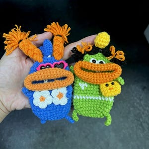 May include: Two handmade crochet character keychains. One is blue with orange accents, sunglasses, and white flower details. The other is green with orange accents and a small yellow pouch. Both have orange hair.