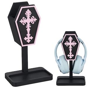 May include: Two black headphone stands with a coffin-shaped design and a pink cross. One stand holds a pair of light blue headphones. The stands have a rectangular base and a vertical support.