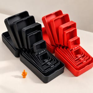 May include: A set of nested rectangular storage boxes in black and red, featuring hinged lids that open to reveal smaller compartments. A small orange figurine depicting a hand with an extended middle finger stands beside the boxes.