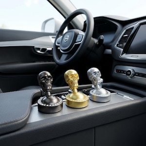 May include: Three skull-shaped car gear shift knobs in black, gold, and silver. Each knob has a detailed skull design and a decorative base. The knobs are displayed inside a car.