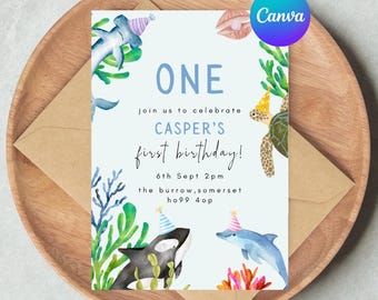 May include: A light blue invitation card with the text "ONE" and "CASPER'S first birthday!" surrounded by watercolor illustrations of marine life, including a shark, orca, dolphin, and turtle. The card is placed on a wooden tray with a brown envelope.