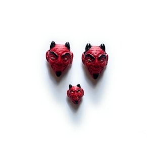 May include: Three red devil head figurines with black horns and beards are arranged on a white background. The devil heads vary in size, with two larger ones and one smaller one. The figurines have a detailed, expressive face.