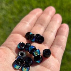 May include: A handful of black, iridescent beads with a hole through the center. The beads reflect a rainbow of colors, including blue, green, and gold. The beads are small and cylindrical, and are likely used for jewelry making.
