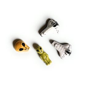 May include: A collection of four small ceramic figurines including a yellow alien head, a small green standing alien figure, and two white space shuttle shapes with black detailing. Each piece features a smooth, glossy finish and distinct shapes.